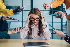 Shot of a stressed out businesswoman surrounded by colleagues needing help. Businesswoman feeling stressed out in a demanding office environment at work. Photo of businesswoman under strain as colleagues request various things from her. Businesswoman feeling overwhelmed by her colleagues in the office. Frustrated businesswoman having a headache while her colleagues are demanding her to work on many things in the office. Stress, anxiety and multitasking businesswoman with headache from workload and laptop deadline in office.